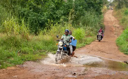 Two people carrying supplies on a motorcycle on a rural road