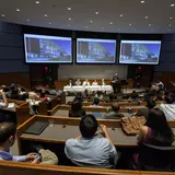 A large lecture hall is filled with attendees seated in tiered rows, facing a panel of five speakers at the front. The panelists are seated at a long table covered with a white tablecloth, and a moderator is standing at a podium to the right. Three large screens above the panel display an image of a modern building. The audience is engaged, with some taking notes and others using laptops or phones. The room is well-lit and designed with wood paneling and modern furnishings, indicating a professional or acad