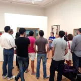 A group of people is gathered in an art gallery, listening to a guide who is explaining a painting on the wall. The attendees are casually dressed and facing the guide, with their backs to the camera. The gallery features several framed paintings on white walls, and the floor is made of light wood. The setting is well-lit, creating a bright and inviting atmosphere.