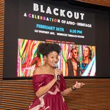 a woman stands with a microphone in front of a digital screen that reads BlackOut