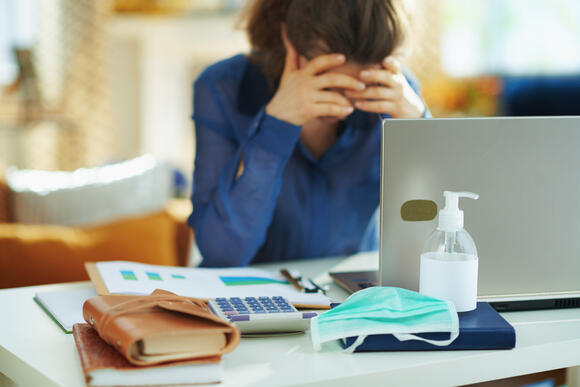 woman at home office desk with head in hands, face mask and hand sanitizer in the foreground