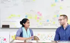 Two colleagues sitting at a table in front of a whiteboard having a discussion