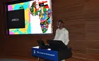 person at a lecturn in front of a screen with image of Africa