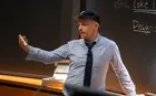 A professor wearing a baseball cap gesturing in front of a chalk board