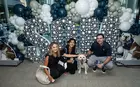 Three people kneeling in front of a balloon display that says “SOM” with a dog