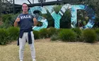 A person wearing a Yale t-shirt and giving a thumbs-up sign stands in front of a large sign that says “SYD”