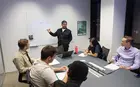 Several people sitting at a table while one standing person talks and gestures at a whiteboard