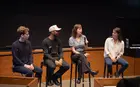 Four people sitting in chairs at the front of an auditorium, with one person speaking into a microphone