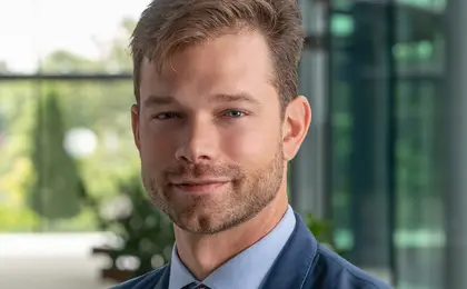 A headshot of a person wearing a suit and tie with a glass wall in the background