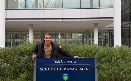A smiling person standing behind an outdoor sign that says “Yale School of Management”