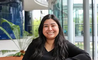 A person with long hair seated on a chair in a light-filled hallway, with a large plant in the background