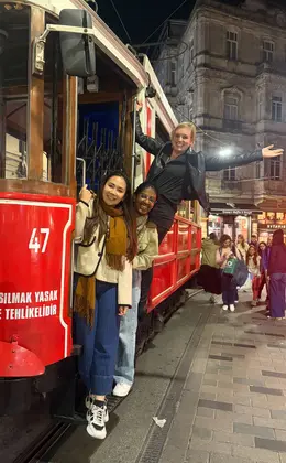 Three people hanging off the side of a tram car in a busy city street at night