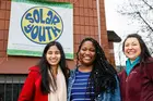 group of three people outside a building with a sign that reads “Solar Youth”