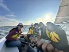 Several people wearing life jackets and sitting on a sailboat, with the ocean in the background