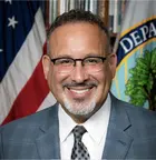 A person wearing glasses and a suit smiling, with an American flag hanging in the background