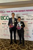 Two people wearing suits standing in front of a conference photo backdrop showing the logos of various corporate sponsors