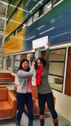 Two women are standing in a modern indoor space with colorful glass panels on the upper floors. One woman is holding up a sign above her head that reads "Welcome" with additional text and imagery, while the other woman points at the sign. Both women are smiling and appear excited. They are dressed casually, with one woman wearing a gray sweater and jeans, and the other wearing a gray top, red scarf, and jeans. The background includes orange seating and glass-walled offices or meeting rooms.