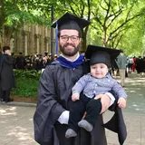 A student in a cap and gown holding a baby, also wearing a graduation cap