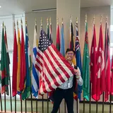 A person standing in front of a row of national flags, holding the American flag