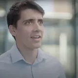 A student wearing a blue button-down shirt speaking in front of a glass wall