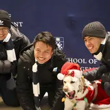 Three people wearing blue and white scarves posing with a dog wearing a Santa hat