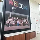 A large wall-mounted poster displaying the word “Welcome” and a group photo of the Asset Management students