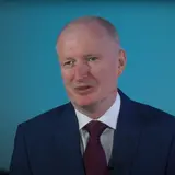 A person wearing a suit and tie, in front of a blue background