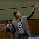 A teacher pointing at a screen, while several students seated in an auditorium listen