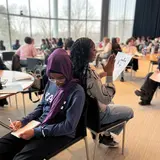 Two high-schoolers sitting back-to-back, each completing a worksheet, in a large room filled with other students