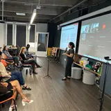 A person giving a PowerPoint presentation in a large room, with other people watching in person and on Zoom