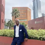 A person with a suit standing on a pedestrian walkway, with several city buildings and two palm trees in the background