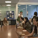 Several people seated at a large conference table, while others walk around the room