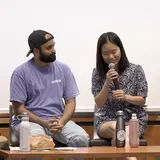 Four people seated in chairs in front of a whiteboard, with one person speaking into a microphone and the others listening