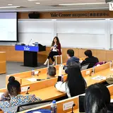 A person seated at the front of a classroom, addressing a group of seated listeners