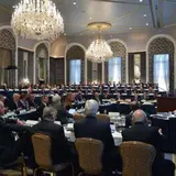 A large conference room is filled with attendees seated at round tables, facing a speaker at a podium. The room is elegantly decorated with chandeliers, high arched windows, and ornate wall designs. The audience is dressed in formal business attire, and the overall atmosphere is professional and sophisticated, suggesting a high-level meeting or event. The room is well-lit with natural light streaming through the windows.