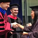 A woman in a graduation cap and gown is shaking hands with a faculty member dressed in academic regalia. The faculty member is wearing a black cap, a red and black gown, and a blue hood. Another faculty member in a black gown and red hood is standing in the background, smiling. The setting appears to be an outdoor or semi-outdoor graduation ceremony, with other graduates and faculty members visible in the background. The moment captures the joyful and celebratory atmosphere of the event.