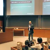 A man is standing at the front of a lecture hall, giving a presentation titled "High Impact Entrepreneurship." He is dressed in business casual attire, wearing a blazer and jeans, and is speaking into a microphone. The presentation slides are projected on screens behind him. The audience is seated in tiered rows, listening attentively, with some people taking notes or looking at the presenter. Several brown paper bags are placed on the desks in front of the audience members, suggesting they may contain refr