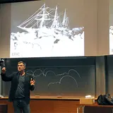 A man is giving a presentation in a lecture hall, standing in front of a projection screen displaying an image of a ship surrounded by ice with the caption "EPIC." He is gesturing with his hands while speaking. Behind him, there are chalkboards with partial drawings and notes. The setting suggests an academic or educational environment, and the presenter is casually dressed in a dark blazer and jeans. The lecture hall features wood paneling and modern design elements.