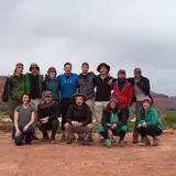 A group of thirteen people is posing for a photo outdoors in a desert landscape. They are dressed in casual outdoor clothing, including jackets, hats, and hiking boots. The group is arranged in two rows, with some people standing and others kneeling in front. Behind them, there are red rock formations and sparse vegetation, suggesting a location in a desert or canyon. The sky is overcast, adding a muted tone to the natural scenery. The group looks cheerful and adventurous, likely on a hiking or camping trip