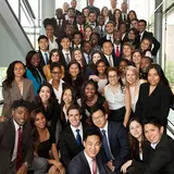 A large group of diverse young professionals is gathered on a staircase for a group photo. They are dressed in business attire, including suits, dresses, and blazers. Everyone is smiling, creating a lively and positive atmosphere. The background features large windows, indicating that they are in a modern, well-lit building. The group appears to be participating in a professional event, workshop, or conference.