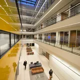 Beckenstein Atrium shown from the second floor, with two people walking on the ground floor. The yellow and blue façade of the atrium is shiny and reflecting the mid-summer night