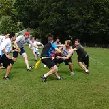 A group of young men is playing a game of flag football on a grassy field. They are actively engaged, with some players reaching for the flags on their opponents' belts. The players are dressed in athletic attire, including shorts, t-shirts, and sneakers. One player in a blue shirt is running towards the goal, while others attempt to block or grab his flag. The background features trees and a glimpse of a house, suggesting a park or recreational area. The atmosphere is energetic and competitive.