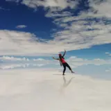 A person is balancing playfully on a vast, reflective surface that mirrors the sky and clouds above. The individual is dressed in casual attire, wearing a pink shirt, dark pants, and a red hat, with arms outstretched as if walking on air. The scene is set against a deep blue sky with scattered clouds, creating a surreal and ethereal atmosphere. The expansive, mirror-like ground blurs the line between earth and sky, enhancing the dreamlike quality of the image.