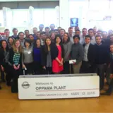 A large group of people is gathered indoors, posing for a photo behind a sign that reads "Welcome to OPPAMA PLANT, NISSAN MOTOR CO., LTD., MAR. 12, 2015." The group includes men and women of diverse backgrounds, all smiling and dressed in business or business casual attire. Behind them, there is a white car and a bright, modern interior, indicating they are at a Nissan facility.