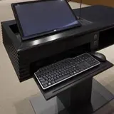 A black lectern is equipped with a computer monitor, keyboard, and mouse. The monitor is tilted and appears to be a touchscreen. A microphone is also attached to the lectern. The background shows rows of seating, indicating the setting is a lecture hall or conference room. The overall setup suggests a space designed for presentations or lectures.